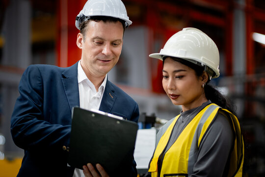Two professional engineer,worker,technician use clipboard discuss work, walk in steel metal manufacture factory plant industry. Black African American man and woman wear hard hat check quality machine