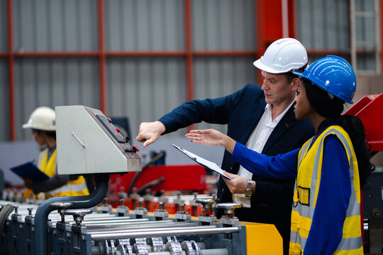 Two professional engineer,worker,technician use clipboard discuss work, walk in steel metal manufacture factory plant industry. Black African American man and woman wear hard hat check quality machine