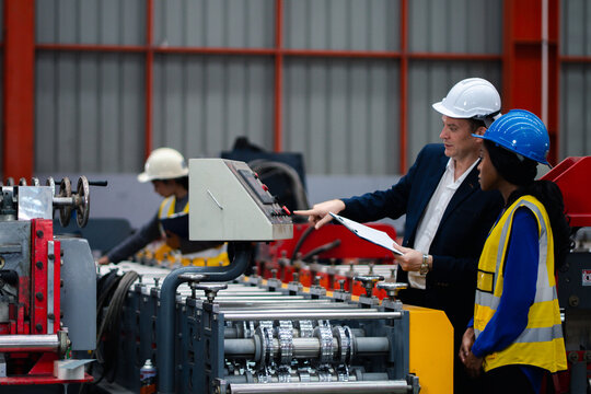 Two professional engineer,worker,technician use clipboard discuss work, walk in steel metal manufacture factory plant industry. Black African American man and woman wear hard hat check quality machine