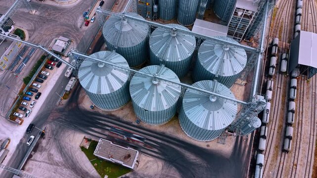 Silver grain silos organized in rows by railway station terminal. Straight down drone perspective of circular steel tanks, asphalt with tire marks, cargo trains on tracks.