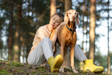 Obraz premium Recreation, building relationships and trust with pet, tranquil woman hiker relaxing in forest with dog, positive female sitting resting head on dogs back enjoying nature, daily walk for mental health