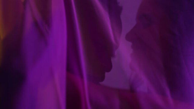 Multiracial couple cuddling under transparent veil in purple light. 
