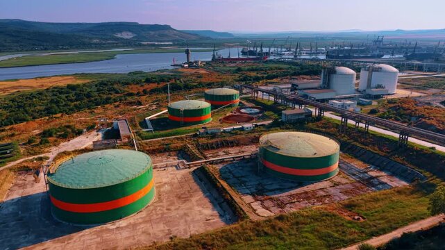 Cylindrical storage tanks near river cargo terminal. An oblique aerial view shows green and orange tanks connected by pipelines, with white silos and harbor cranes behind.