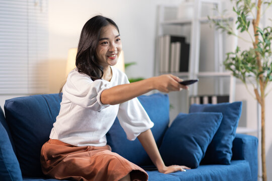 Happy young Asian woman watching TV at home. Young Asian woman smiling, holding a remote control, sitting on a blue couch in her living room, enjoying entertainment.