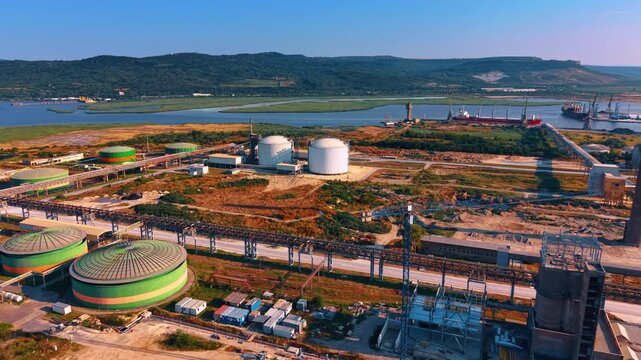 Coastal industrial plant with gas reservoirs near water channel. White spherical containers and round colorful tanks stand on a territory bordered by a river and hills.
