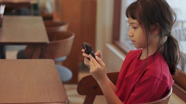 Asian kid using a smartphone at restaurant. Cute Thai girl excitedly watching video or playing games. Shot with selective focus and beautiful copy space for text. Childhood digital lifestyle concept.