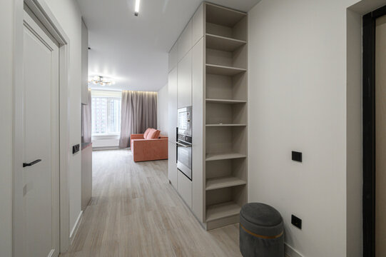 sleek hallway features a built‑in wardrobe with open shelves, light wood flooring, gray upholstered ottoman, and minimalist white doors