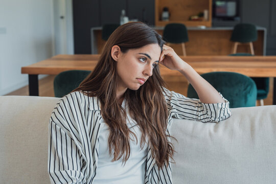 Young woman sitting on a comfortable sofa in her living room, experiencing feelings of sadness, loneliness, and reflection while looking away in thought