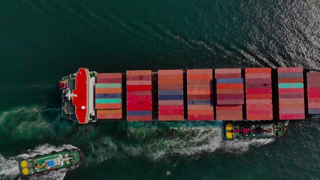 Large red container ship powerfully driven through dark ocean waters by tugboat and small support boat, generating dynamic white wave patterns during nighttime maritime transport.