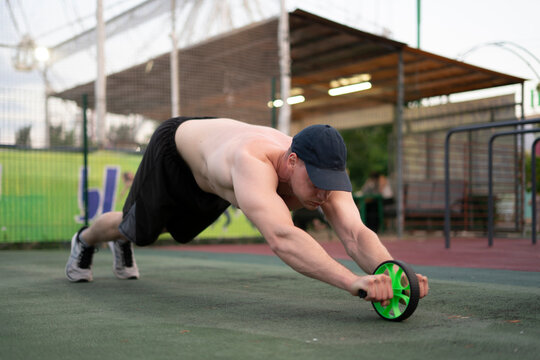 A man performs an ab wheel exercise on a green surface in an outdoor gym late in the day with others in the background