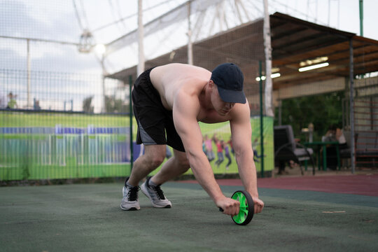 A man trains using a fitness wheel on a court while others watch from nearby. It is late afternoon and sunny