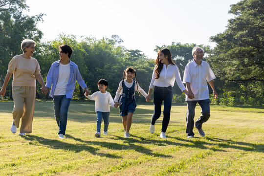 Happy multi-generational Asian family holding hands and walking together in a sunny green park. Grandparents, parents, and kids enjoying outdoor leisure time, promoting love and bonding
