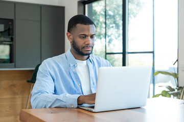 Fototapeta premium Young man seated at a wooden table using a laptop in a bright modern home office, focused on remote work and productivity with natural light from large windows and a casual, professional look.