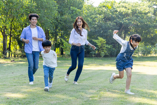 Happy Asian family with parents and young children running together on a green lawn in a sunny park. Enjoying outdoor activities, a healthy lifestyle, and quality time during summer leisure