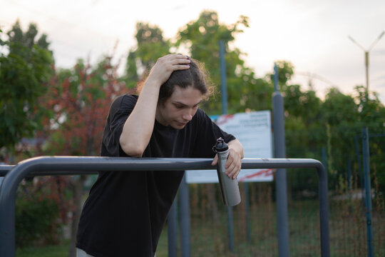 A young man takes a break after exercising in a park, holding a water bottle while watching the surroundings
