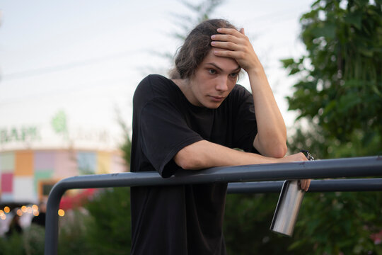 A young person is leaning on a railing with a worried expression in a park at dusk near some trees and buildings