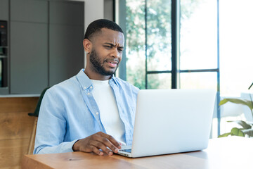 Fototapeta premium Young black man looking confused and upset at laptop screen, experiencing technology issues or a problem working from home in his modern kitchen