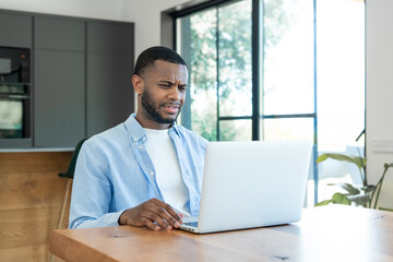 Fototapeta premium A young man working from home looks frustrated and confused while using his laptop in a bright modern home office, conveying remote work stress, technical issues and concentration under pressure.