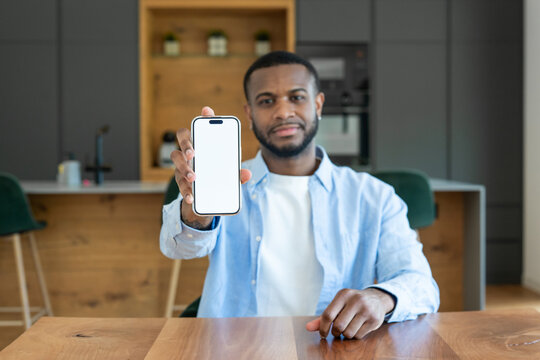 A young man sits at a wooden kitchen table holding a smartphone toward the camera with a blank white screen, suggesting app demo, mobile marketing, remote work, communication and modern lifestyle.