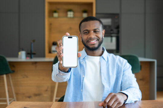 Smiling man seated in a modern kitchen holds a smartphone toward the camera with a blank white screen, perfect for app mockups, tech presentations, social media promotion, and remote