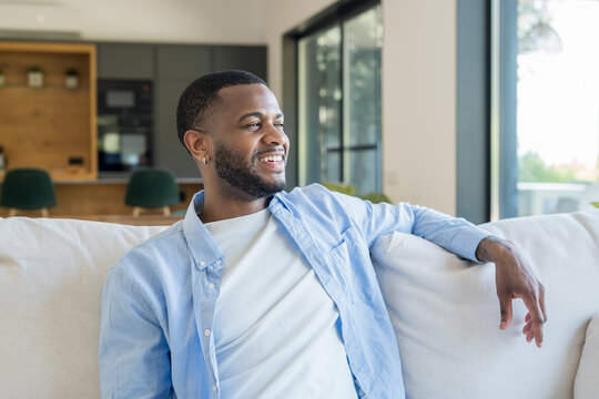 Smiling young man relaxes on a comfortable sofa in a bright, open-plan living room. Natural light fills the space, conveying calm, contentment and casual everyday home lifestyle.