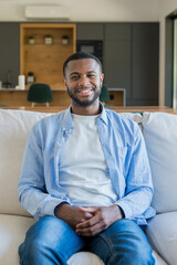 Fototapeta premium Young black man smiling at camera while sitting comfortably on a sofa in a modern home, representing relaxation, happiness, and lifestyle in a domestic indoor setting