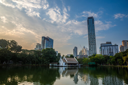 Scenic view of Lumphini Park in Bangkok featuring a lake with reflection of city skyscrapers and pavilion under cloudy sky