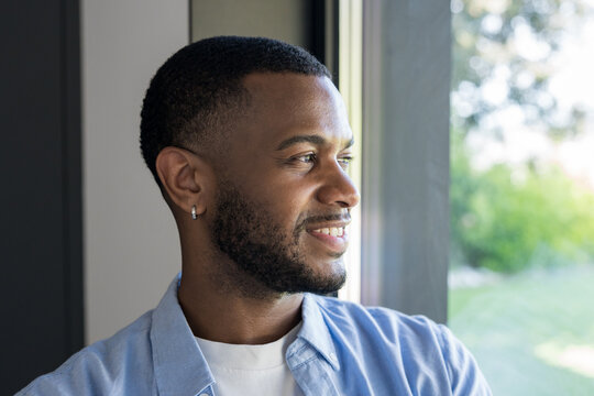 Happy young black man looking away with a hopeful expression and smiling while standing near a bright window, reflecting concepts of contentment and future aspirations