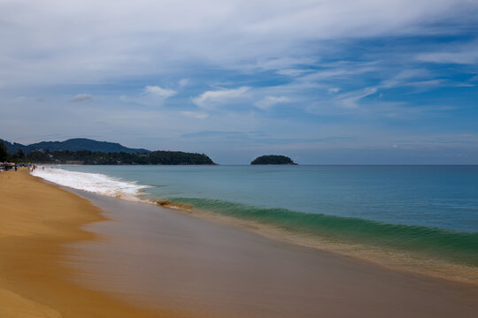 Tropical beach view with incoming waves on the sand and a lush green island coastline under cloudy sky