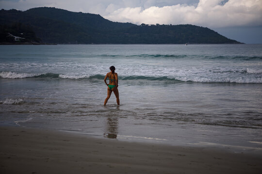 Young female traveler in swimsuit and hat looking back on a beautiful turquoise tropical sea beach with blowing blue cape
