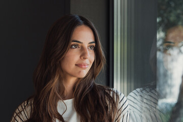 Fototapeta premium Young brunette woman standing beside a window, looking out with a thoughtful and serene expression, her reflection visible on the glass, conveying a sense of peace and contemplation indoors