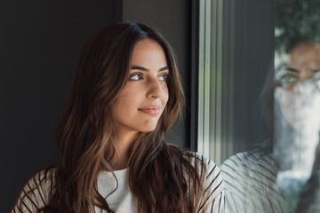 Fototapeta premium Thoughtful young woman gazes out a window with her soft reflection on the glass, a natural lifestyle portrait that conveys calm, introspection, peaceful mood and quiet morning light.