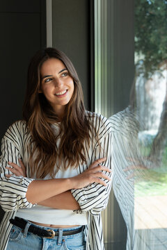 Young woman smiling, standing by a window, arms crossed, and looking away with natural light reflecting on the glass