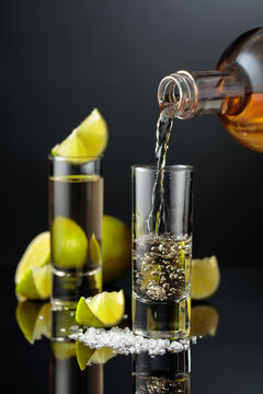 Tequila shots with salt and lime slices on a black reflective background.
