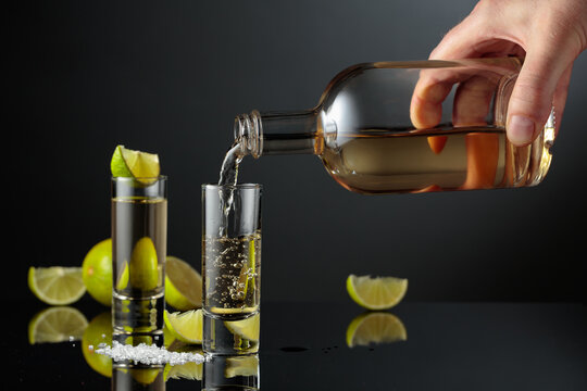 Tequila shots with salt and lime slices on a black reflective background.