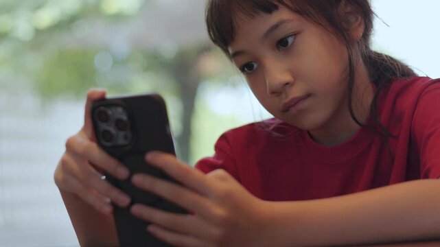 Close up Asian kid seriously focusing on a smartphone at a restaurant. Thai girl intently playing mobile games or watching content. Shot with selective focus and copy space. Digital lifestyle concept.