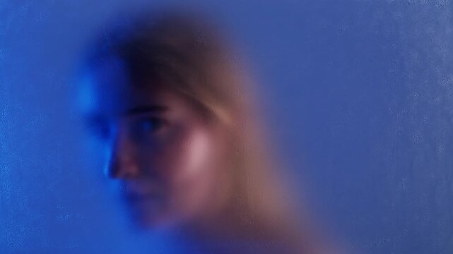 Woman looking at camera through wet matte glass in blue light. She is feeling sad and in need of help with depression