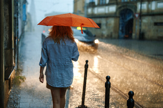 Woman walking with orange umbrella on sidewalk in heavy rain. City street during rain storm with dramatic weather and glowing reflections
