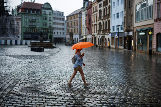 Woman crossing town square with orange umbrella in rain. Female person walking across wet cobblestones in historic city during stormy weather