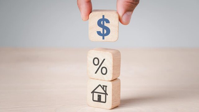 Close-up of a hand stacking wooden blocks with house, percentage, and dollar sign icons, representing mortgage interest rates, real estate investment, home loan property finance, and banking concept.