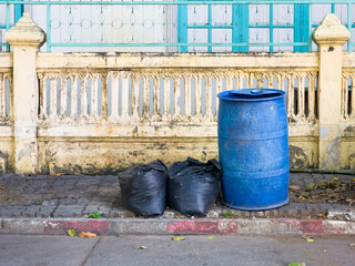 Naklejka premium Two black garbage bags and a blue plastic barrel placed on a sidewalk beside a street in front of an old decorative wall.