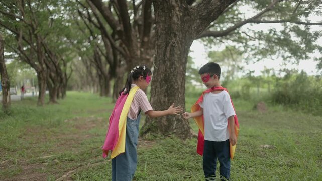 Two adorable Asian children wearing colorful superhero capes standing close together on lush green grass under large shady trees, interacting playfully during outdoor leisure.