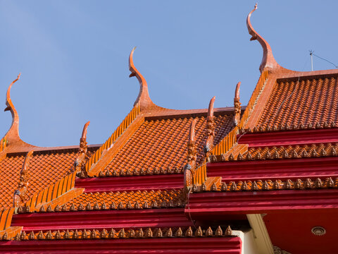 Traditional Thai temple roof featuring layered orange and red tiles with decorative chofah finials against a blue sky.