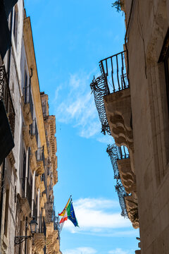 a Sicilian alleyway lined with ancient palaces featuring typical Baroque balconies, with colourful flags hanging from the wall in the background, set against an intensely blue sky