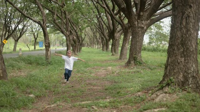 Active young Asian boy dressed in a clean white t-shirt running joyfully down a beautiful forest pathway under lush green trees in a sunny public nature park during summer season.