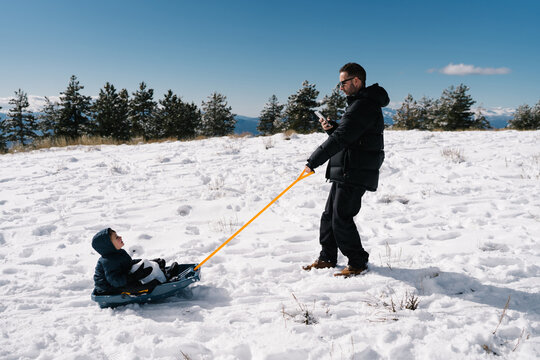 Father pulling son on sled while using smartphone