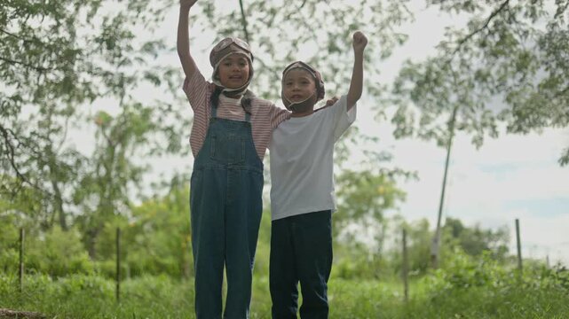 Two adorable Asian kids wearing cute aviator pilot hats standing on green grass field, raising their arms high with pure excitement and happy smiles under beautiful shady trees.