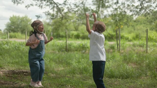 Active little Asian boy and girl wearing pilot hats standing together on fresh green grass field, looking up and pointing towards the sky with great imagination in nature park.
