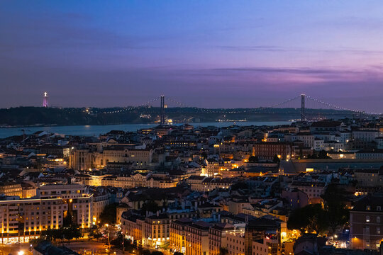 High angle panoramic view of Lisbon city skyline at dusk with illuminated streets, historic architecture the Tagus river and the suspension bridge