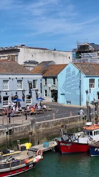Weymouth Dorset England UK. 08.07.2026. Video, Boats bob gently in the water next to a waterfront street lined with colourful buildings and people enjoying outdoor seating